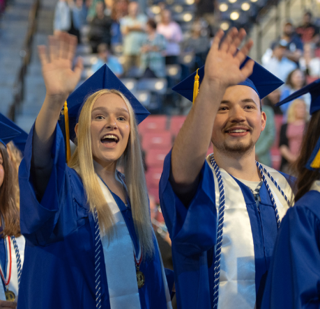 Two students wearing caps and gowns at a graduation ceremony and waving