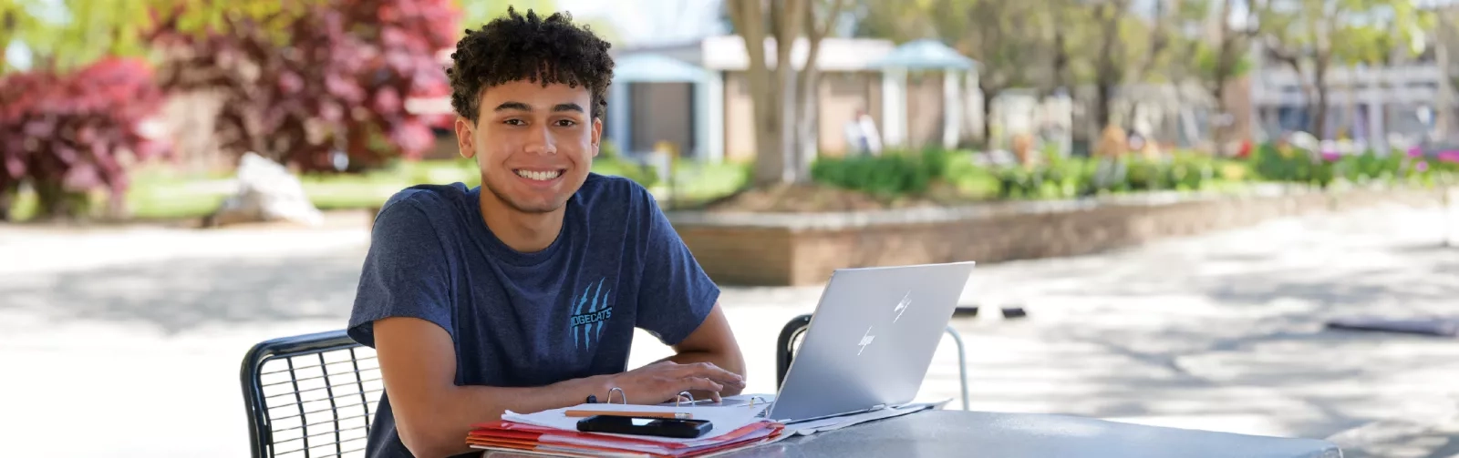 A student wearing a Ridgecats shirt seated outside at a table with a laptop.