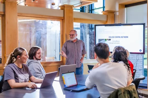 A man standing in a small conference room beside a TV screen with four people seated around a table looking at laptops.